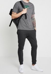 Grå t-shirt med grön camouflage-logotyp, tillsammans med mörka joggers och vita sneakers. Svart ryggsäck slängd över axeln, vilket visar en tatuering på armen.