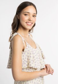 Top cu imprimeu floral în galben deschis, cu mâneci volan. Prezintă un decolteu rotund și o croială relaxată. Accentele includ flori mici de culoare mov.