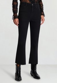 Jeans neri a zampa con orlo sfrangiato, realizzati in denim. Abbinati a una maglia nera trasparente e stivaletti neri alla caviglia.