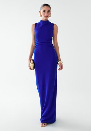 Robe bleue longue jusqu'au sol avec un col haut, un corsage ajusté et une texture lisse. Le mannequin tient une pochette dorée et porte de grandes boucles d'oreilles en or.