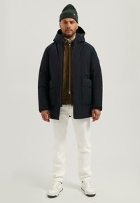 Plyšová bunda tmavě modré barvy s kapucí, se dvěma předními kapsami, nošená přes hnědou semišovou bundu, v kombinaci s bílými kalhotami a teniskami.