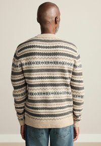 Béžový pletený sveter s geometrickým vzorom v tmavosivej a čiernej farbe, rebrované manžety a okrúhly výstrih. Nosený s modrými rifľami.