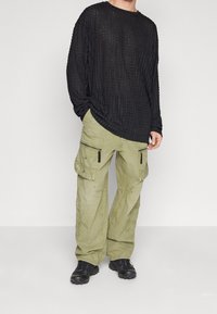 Person wearing black textured long-sleeve shirt, loose olive cargo pants with zippers and pockets, and black athletic shoes, standing against white background.