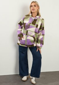 Fleece pullover in een patchworkpatroon van paars, groen en ivoor. Voorzien van een halve rits, hoge kraag en een zak aan de voorkant. Gekocht met een brede jeans.