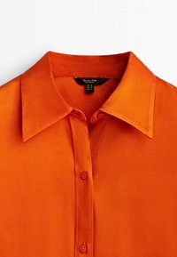 Chemise orange vif à col avec patte de boutonnage, texture lisse et étiquette de marque visible à l'intérieur de l'encolure.