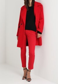 Vrouw in een rode jas en broek met een zwarte gestructureerde trui en zwarte hoge hakken, staande tegen een effen achtergrond.