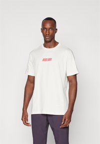 Tricou din bumbac alb-beige, cu guler rotund și mâneci scurte, având inscripția roșie "Riding Hoops" pe piept, asociat cu pantaloni mov.