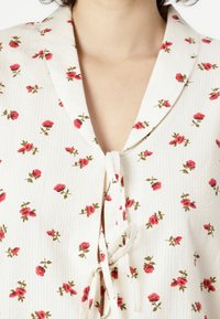 Bluse med blomsterdesign i lys krem med røde roser og grønne blader. Har en myk tekstur, krage og en knyttet åpning foran.
