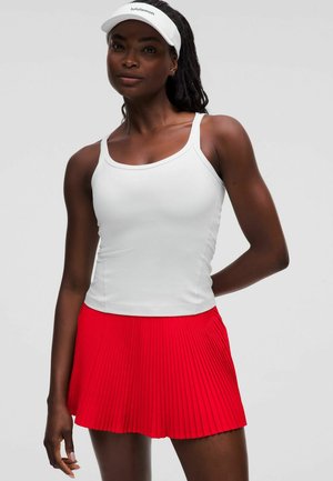 Donna atletica con canottiera bianca, gonna plissettata rossa e visiera bianca Lululemon, in piedi con una mano dietro la schiena su sfondo grigio.