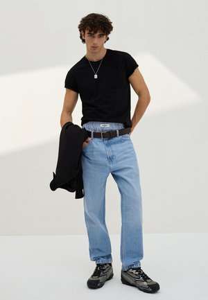 Mladý muž v čiernom tričku, modrých džínsoch, čiernom opasku, sivých teniskách a s čiernou bundou v ruke, stojaci proti jednoliatemu svetlému pozadiu.