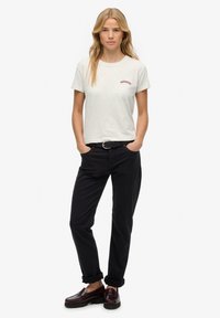 Camiseta de manga corta color gris claro con bordado rojo de "Superdry", combinada con pantalones negros y mocasines burdeos. Ajuste casual, diseño relajado.