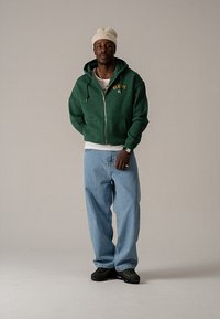 Zelená mikina na zips so žltým textom "BLK IVS NY", doplnená voľnými modrými džínsami a čiernymi teniskami. Outfit dotvára béžová čiapka.