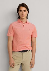 Polo-shirt en coton à manches courtes de couleur corail avec un col et trois boutons, porté avec un pantalon kaki et une ceinture noire. Texture subtilement visible.