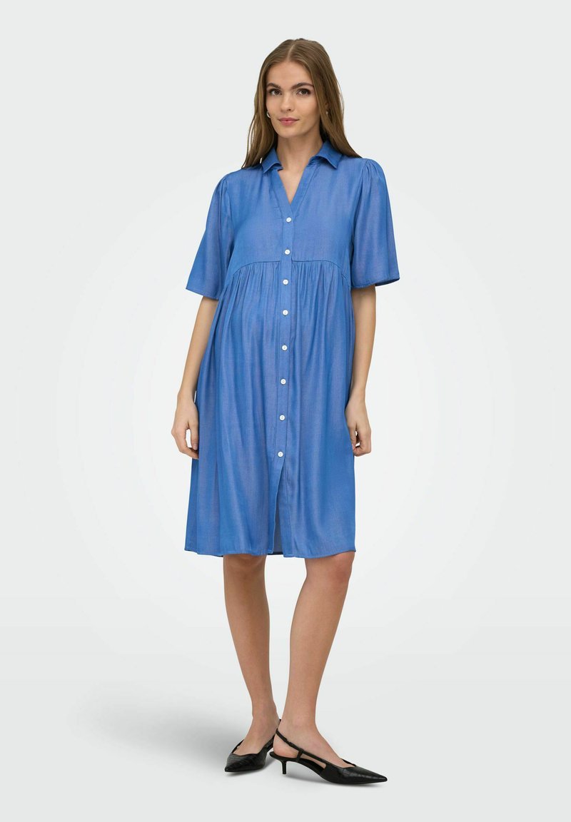 Femme portant une robe bleue boutonnée jusqu'aux genoux, manches courtes, col, avec des escarpins à talons bas slingback noirs, sur un fond uni.