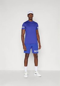 Homem a usar uma camisa de manga curta azul e calções com texto branco, boné branco, meias brancas e ténis brancos, de pé contra um fundo simples.