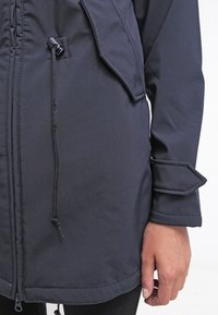 Close-up van een hand naast een donkerblauwe jas met een ritssluiting aan de voorkant, een trekkoord, een klepzak en een manchetband met stiksels.