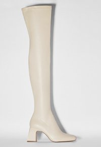 Botte haute en cuir couleur crème, jusqu'au genou, avec bout arrondi et talon bloc sur fond clair uni.