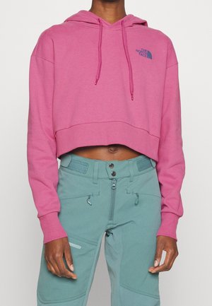 Felpa cropped in cotone rosa brillante, con un logo frontale in blu navy, cappuccio con cordino e polsini a coste. Indossata con pantaloni color teal.