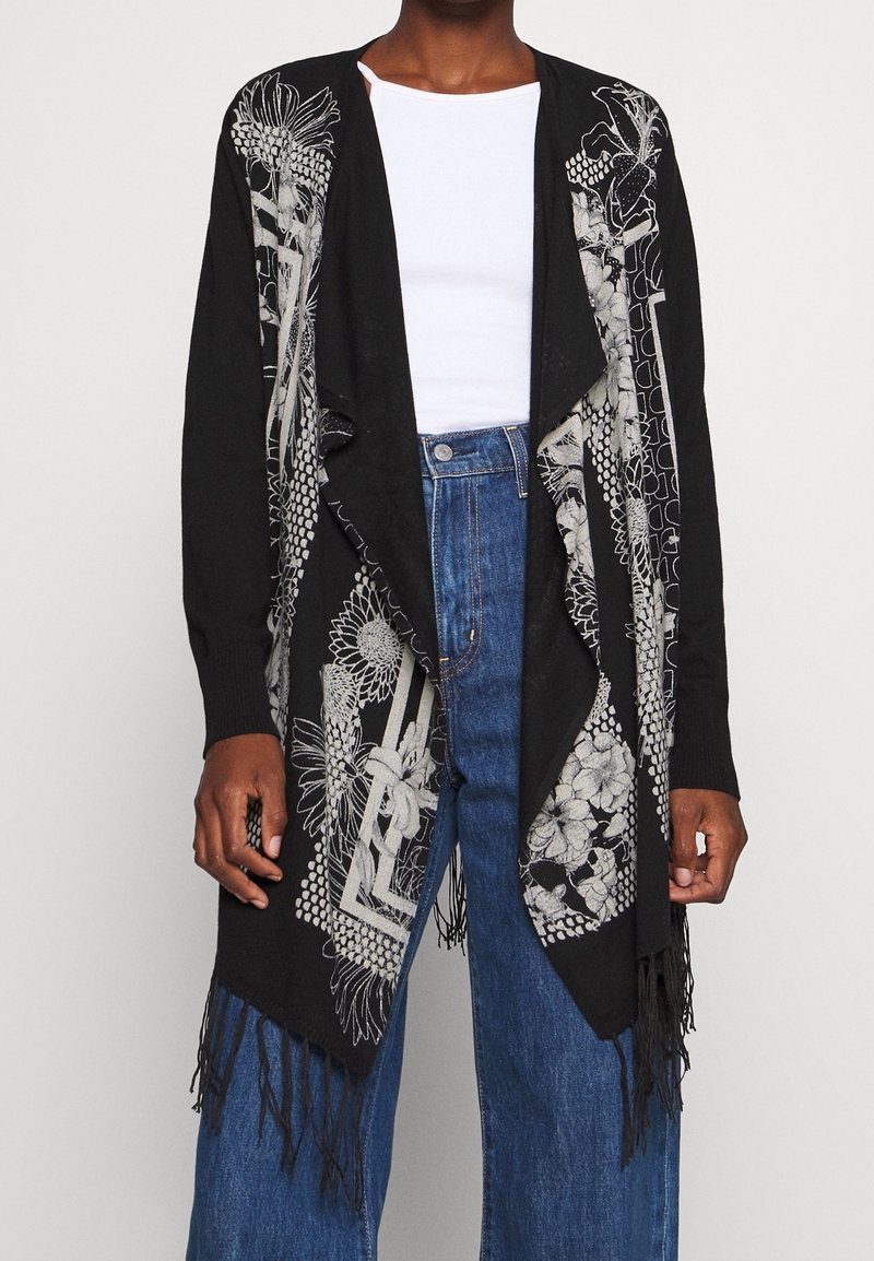 Svart kofta med blom- och geometriskt silverbroderi, öppen fram och fransig nederkant. Buren över en vit top och blå denim.