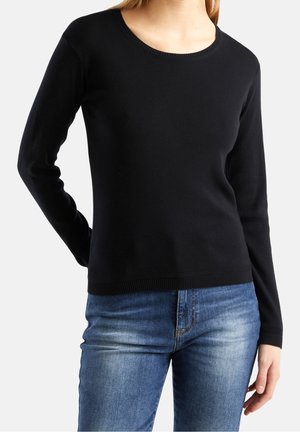 Femme portant un pull noir ajusté à manches longues associé à un jean bleu délavé moyen, debout devant un fond blanc uni.