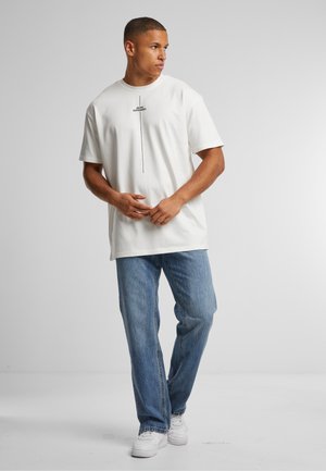 T-shirt en coton blanc avec un graphique sur le devant, associé à un jean en denim bleu clair et des baskets blanches, sur un fond neutre.