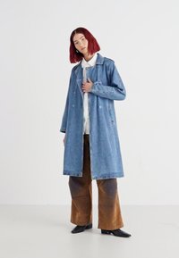 Cappotto trench in denim di un azzurro chiaro, design doppiopetto e cuciture a vista, abbinato a pantaloni palazzo marroni.