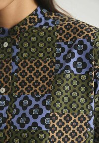 Chemise à boutons avec un design en patchwork aux couleurs bleue, verte, marron et noire ; motifs floraux avec divers accents. Tissu lisse.