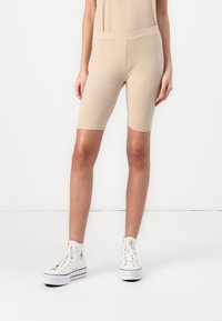 Beige cyklistické kraťasy vyrobené z měkkého, pružného materiálu. Vyznačují se vysokým pasem a přiléhavým designem, dosahujícím do poloviny stehen. Kombinované s bílými teniskami.