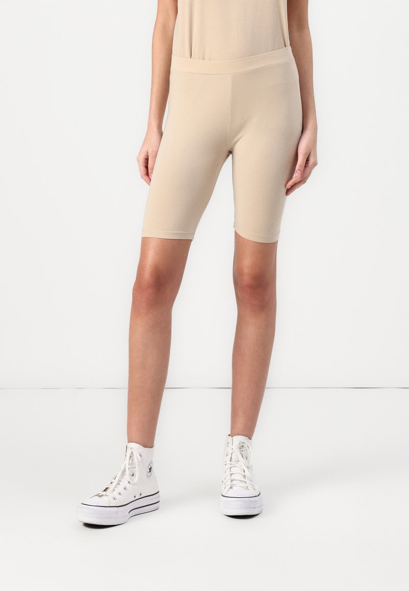 Beige cyklistické kraťasy vyrobené z měkkého, pružného materiálu. Vyznačují se vysokým pasem a přiléhavým designem, dosahujícím do poloviny stehen. Kombinované s bílými teniskami.