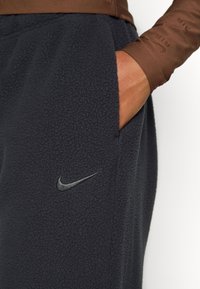 Joggingi negri din fleece cu buzunare laterale, având o textură ușor periată și un logo metalic Nike aproape de buzunar.