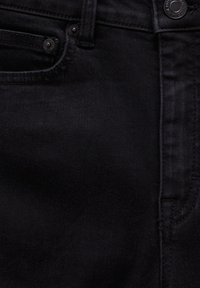 Gros plan sur un jean en denim noir montrant la poche avant, le bouton, la passants de ceinture et les détails de couture.