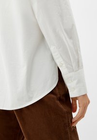 Witte, gekraagde blouse met een gestructureerde afwerking, voorzien van knoopmanchetten. Gecombineerd met een paar bruine corduroy broeken, die een zachte, geribde textuur vertonen.