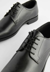 Zapatos de vestir de cuero negro con un acabado suave, diseño de cordones, punta redonda y suela plana. Costuras visibles y acentos minimalistas.