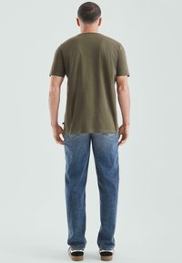 Krótki rękaw oliwkowozielonej koszulki, standardowy krój, w połączeniu z jasnoniebieskimi dżinsami. Osoba stoi na neutralnym tle.