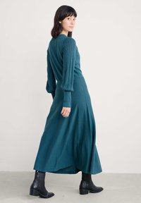 Žalioji suknelė ilgomis rankovėmis su tekstūruotomis pūstomis rankovėmis, prigludusia liemene ir laisvai krentančia sijona. Dėvima su juodais auliniais batais iki kulkšnies. Lygaus audinio apdaila.