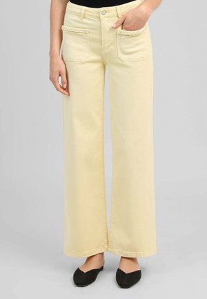Femme portant un jean taille haute à jambes larges de couleur jaune pâle, avec des poches plaquées devant et des ballerines noires pointues sur fond gris.