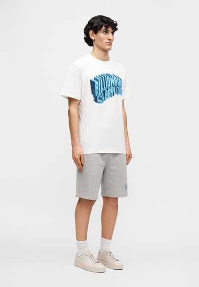 Λευκό βαμβακερό t-shirt με μπλε τύπωμα, συνδυασμένο με γκρι σορτς. Το μοντέλο φοράει μπεζ αθλητικά παπούτσια και λευκές κάλτσες. Καθαρός σχεδιασμός, casual εφαρμογή.