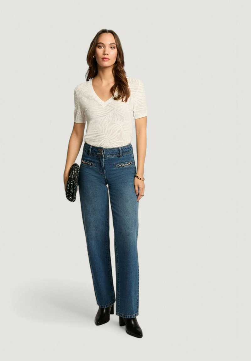 Top bianco a maniche corte con scollatura a V e motivo in rilievo a foglie, abbinato a jeans blu a gamba dritta con vita alta e stivaletti neri con tacco.