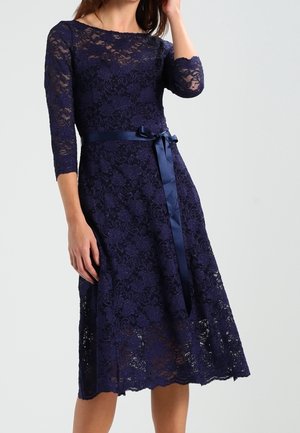 Granatowa koronkowa sukienka z rękawami do połowy przedramienia, dopasowaną górą i rozkloszowanym dołem. Ozdobiona satynowym paskiem w talii i falowanym brzegiem sukienki.