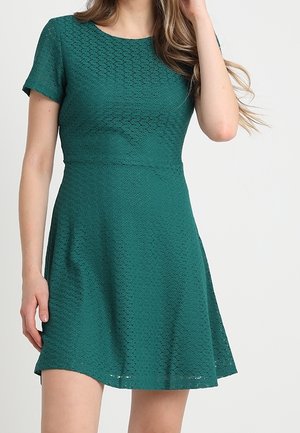 Donna che indossa un abito verde teal a maniche corte con trama, vita aderente e gonna svasata, in piedi davanti a uno sfondo chiaro e semplice.