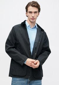 Young man wearing dark zip-up jacket over light blue sweater and shirt, fastening jacket, standing against plain white background.