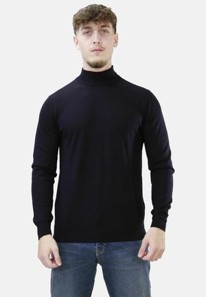 Giovane uomo con capelli castano chiaro che indossa un maglione nero a collo alto e jeans blu su uno sfondo chiaro uniforme.