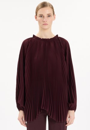 Donna che indossa una blusa plissettata a maniche lunghe di colore viola scuro con scollo arricciato, in piedi davanti a uno sfondo bianco semplice.