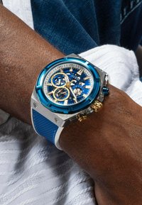 Blaues und silbernes Chronographen-Armbanduhr mit einem strukturierten blauen Armband, goldenen Akzenten und einem detaillierten mehrschichtigen Zifferblatt, das mehrere Unterzifferblätter anzeigt.