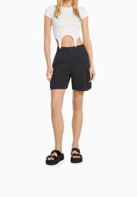 Žena v bílém krátkém topu s výřezy a černých cargo šortkách, doplněná černými platformovými sandály, stojící na bílém pozadí.