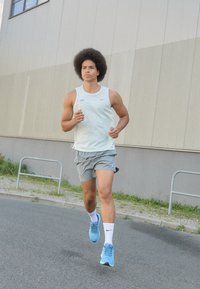 Camiseta atlética sin mangas de color azul claro y pantalones cortos grises, corriendo sobre el pavimento con zapatillas deportivas azules. El fondo incluye un edificio gris y vegetación.