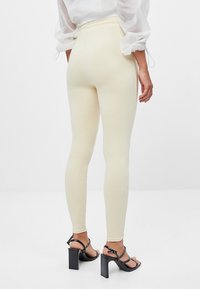 Osoba v krémově zbarvených priléhavých legínách, bílé topu s nafouknutými rukávy a černých sandálech na vysokém podpatku stojí na jednobarevném bílém pozadí.