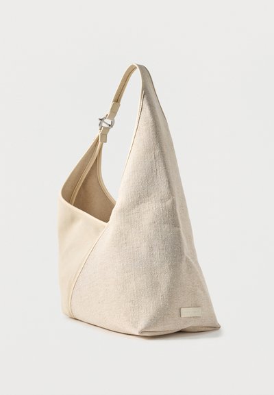 Sac à bandoulière en toile beige, avec une large sangle incurvée, une boucle en métal et une petite étiquette à l’avant, présenté sur un fond clair uni.