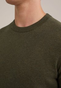 Sötétzöld kötött pulóver kerek nyakkal, sima textúrával és bordázott nyakkivágással, a szövet közeli képe látható.