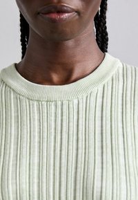 Zbliżenie osoby noszącej jasną zieloną sweter z prążkami, ukazujące dolną część twarzy, szyję i warkocze.